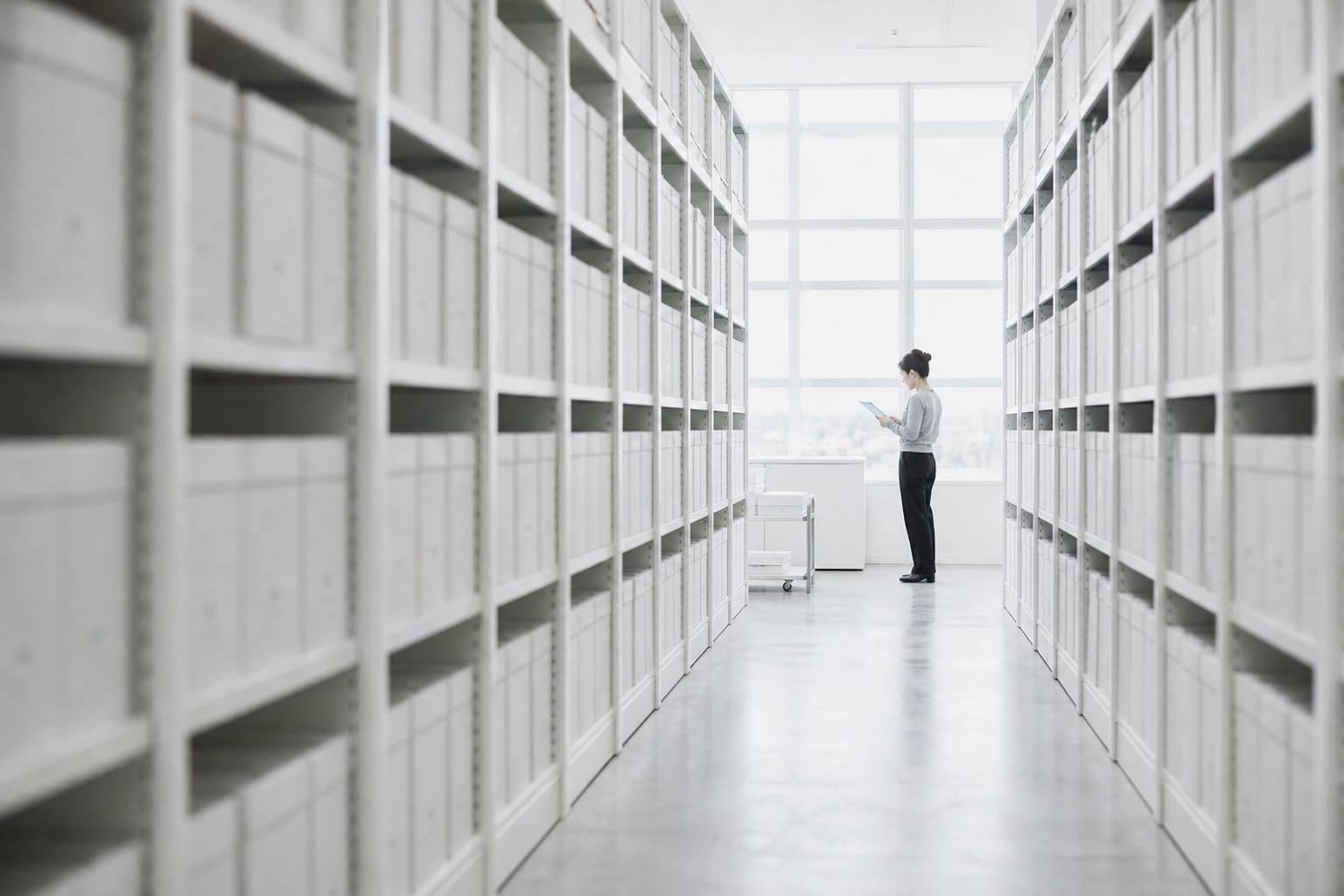 Archivo con estanterías blancas llenas de cajas iguales y una persona al fondo del pasillo